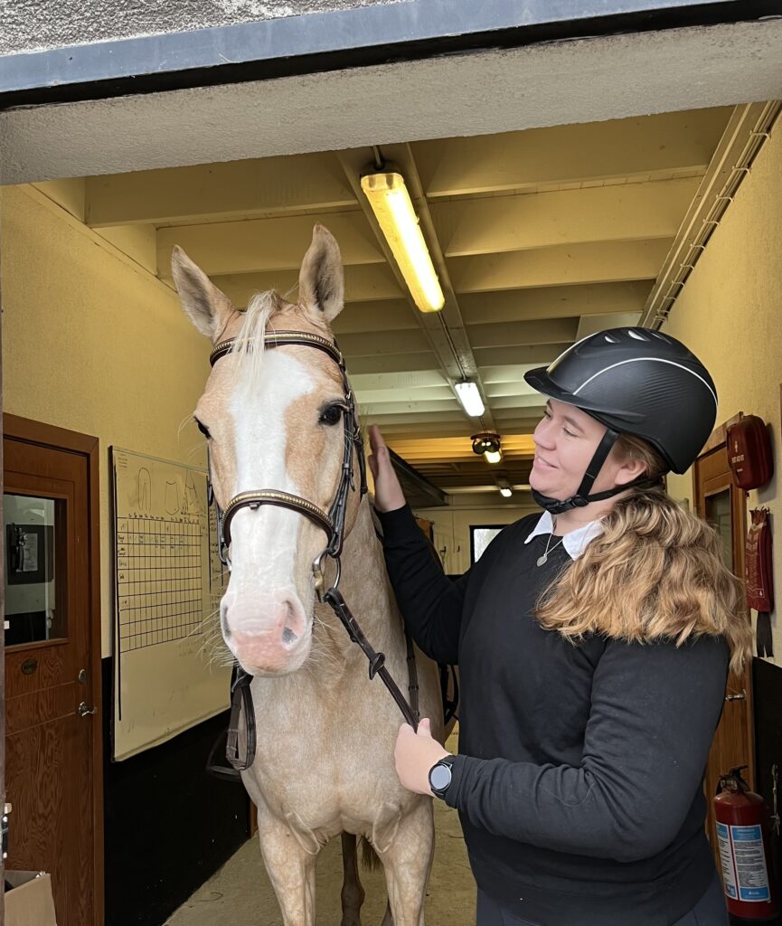 Louise Sundling tillsammans med en isabellfärgadhäst i en stallgång, utrustade för en ridtur. Louise klappar hästen och ser glad ut. 
